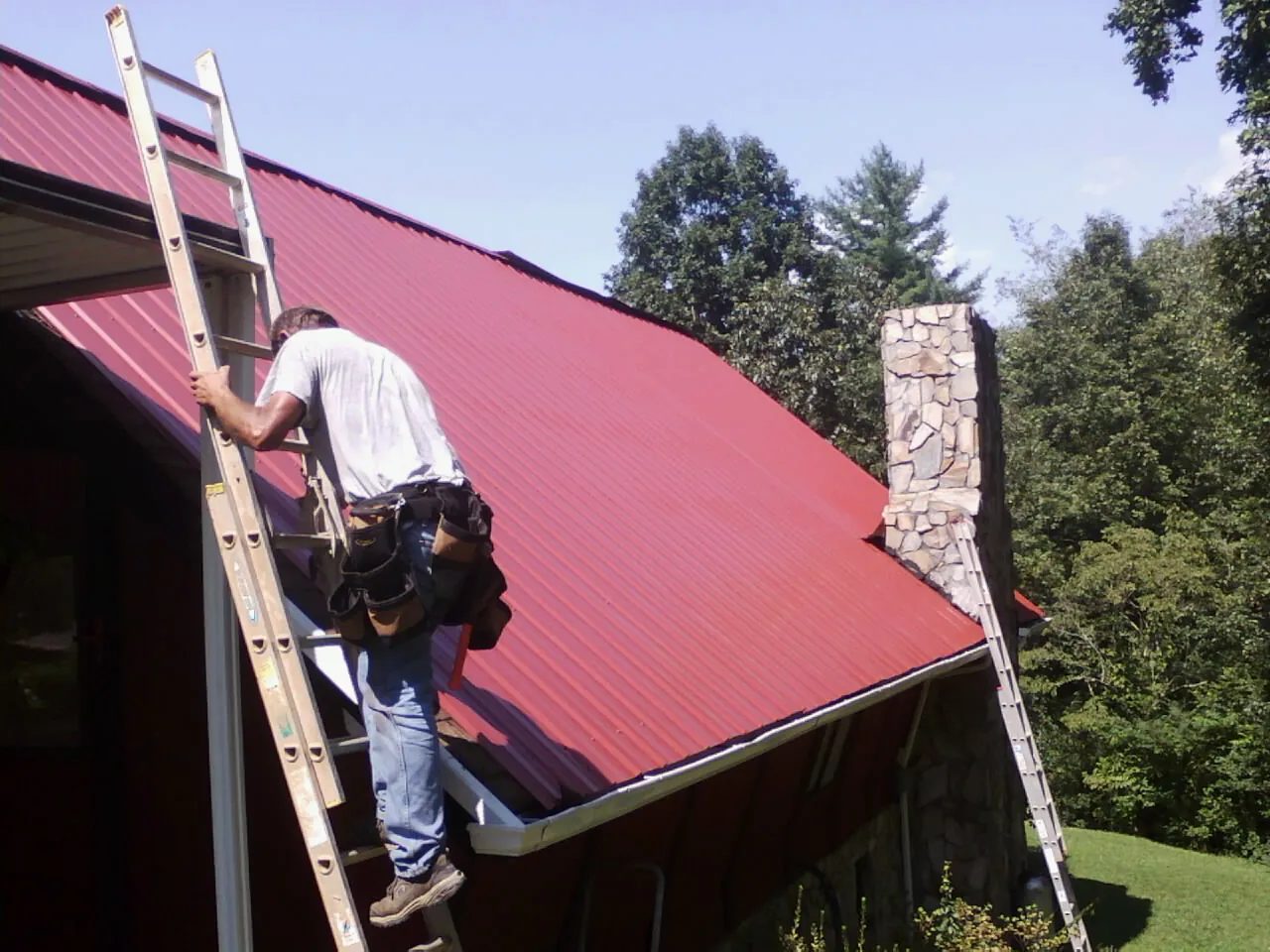 Roofer on a ladder beside a red metal roof and stone chimney — Soffit & Fascia Repair by Roofing Brothers in Rathdrum